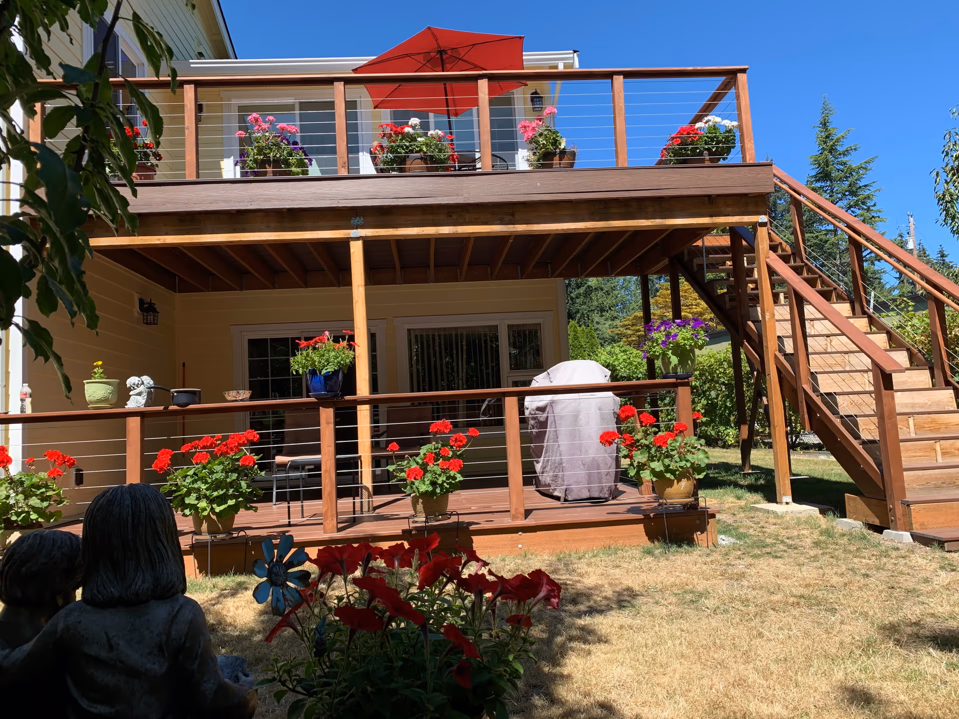 Backyard view of a two-story house with a wooden deck and stairs leading to the upper level. The deck is decorated with multiple flower pots containing red and pink flowers, and a red patio umbrella is open on the upper deck. There is a covered grill on the lower deck, and a small statue of two children sitting is visible in the foreground among flowers. The sky is clear and blue.