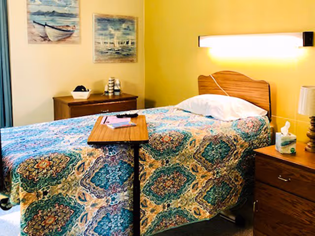 A single bed in a healthcare facility room with a colorful patterned bedspread. The bed has a wooden headboard and a white pillow. A small overbed table is positioned on the bed with a remote control and a book on it. There are two wooden nightstands on either side of the bed, one with a lamp and a box of tissues. The walls are painted yellow and decorated with two framed paintings of boats on water.