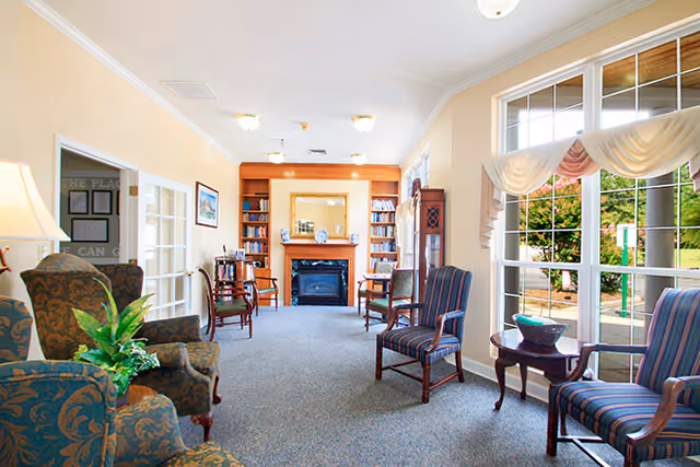Well-lit sitting room with upholstered chairs, a fireplace flanked by bookshelves, and large windows overlooking greenery.