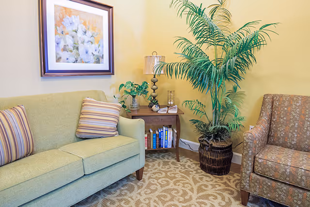 A cozy living room corner with a light green sofa adorned with striped pillows, a wooden side table holding a lamp, a small plant, and books on the lower shelf. A large potted palm plant is placed next to the table, and a patterned armchair is partially visible. The walls are painted yellow and decorated with a framed floral painting. The floor is covered with a beige carpet featuring a swirling pattern.