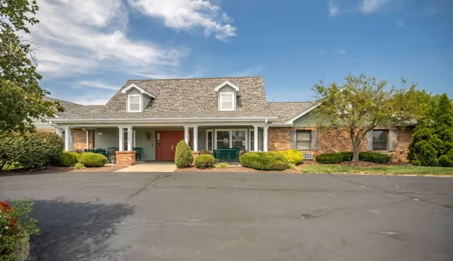 Front exterior view of a single-story brick building with a covered porch, two dormer windows on the roof, and a paved driveway in front. The building is surrounded by neatly trimmed bushes and trees under a partly cloudy sky.
