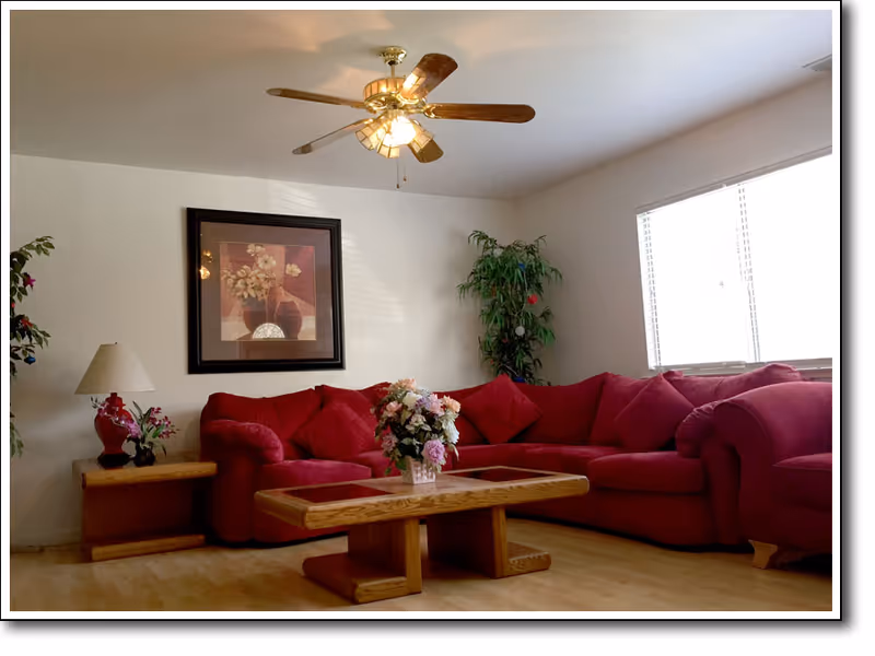 A cozy living room with a large red sectional sofa adorned with matching red cushions. A wooden coffee table with a floral centerpiece sits in front of the sofa. There is a wooden side table with a lamp and a small plant on it. A framed floral painting hangs on the white wall above the sofa. A ceiling fan with lights is mounted on the ceiling. A large window with blinds allows natural light to brighten the room. A tall green plant is placed in the corner near the window.