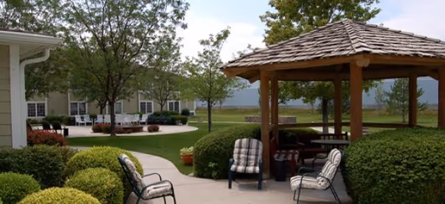 Outdoor seating area with cushioned chairs arranged around a wooden gazebo with a shingled roof. The area is surrounded by green bushes, trees, and a paved walkway. In the background, there is a building with windows and more outdoor seating under trees.