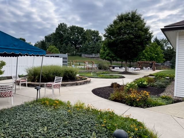 Outdoor garden area at The Terraces at Capitol Village featuring paved walkways, green shrubs, flowering plants, several white chairs with patterned cushions, a blue canopy, and large trees under a cloudy sky.