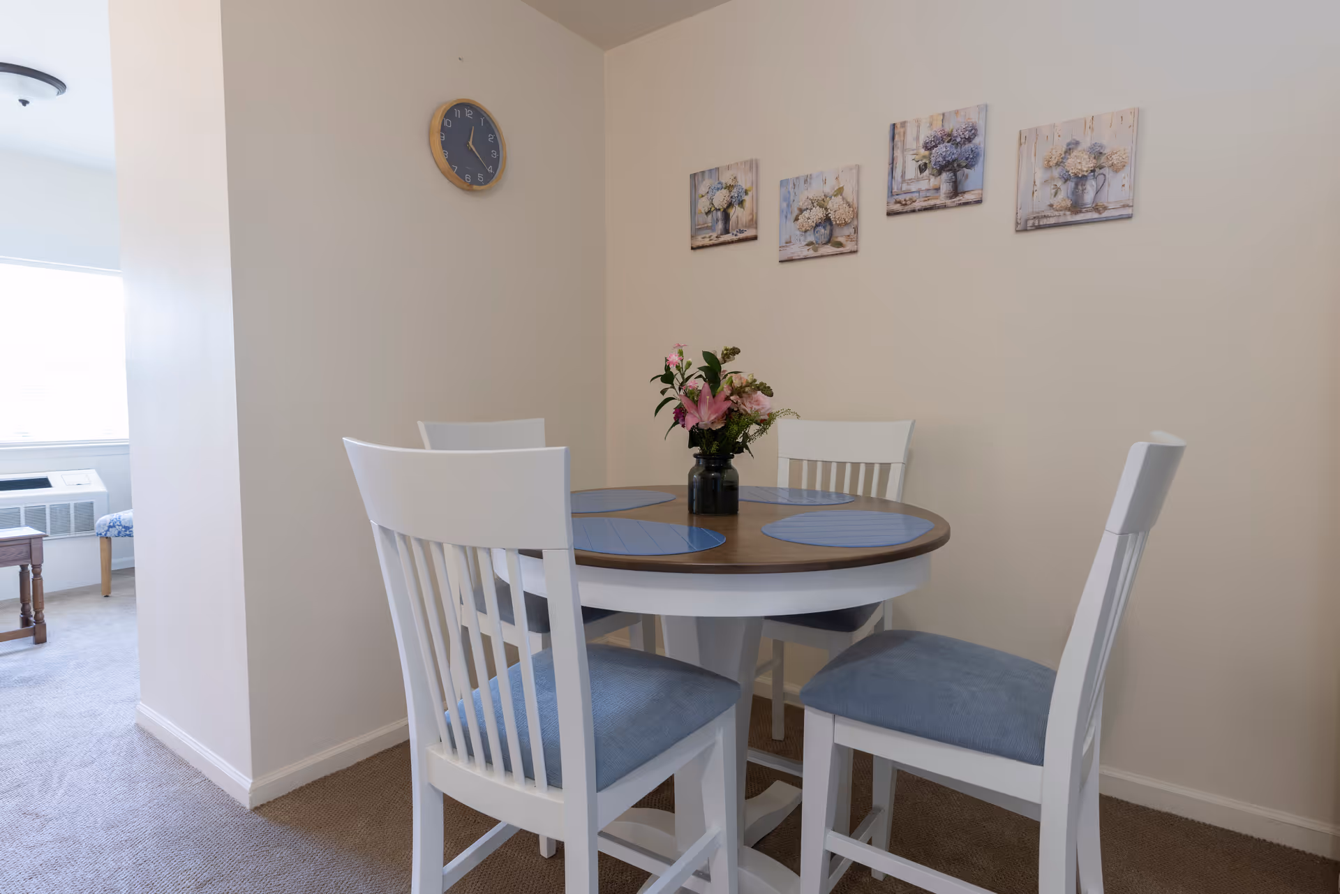 Small dining area with a round table, four white chairs with blue cushions, blue placemats and a vase of flowers beneath a wall clock and floral prints.