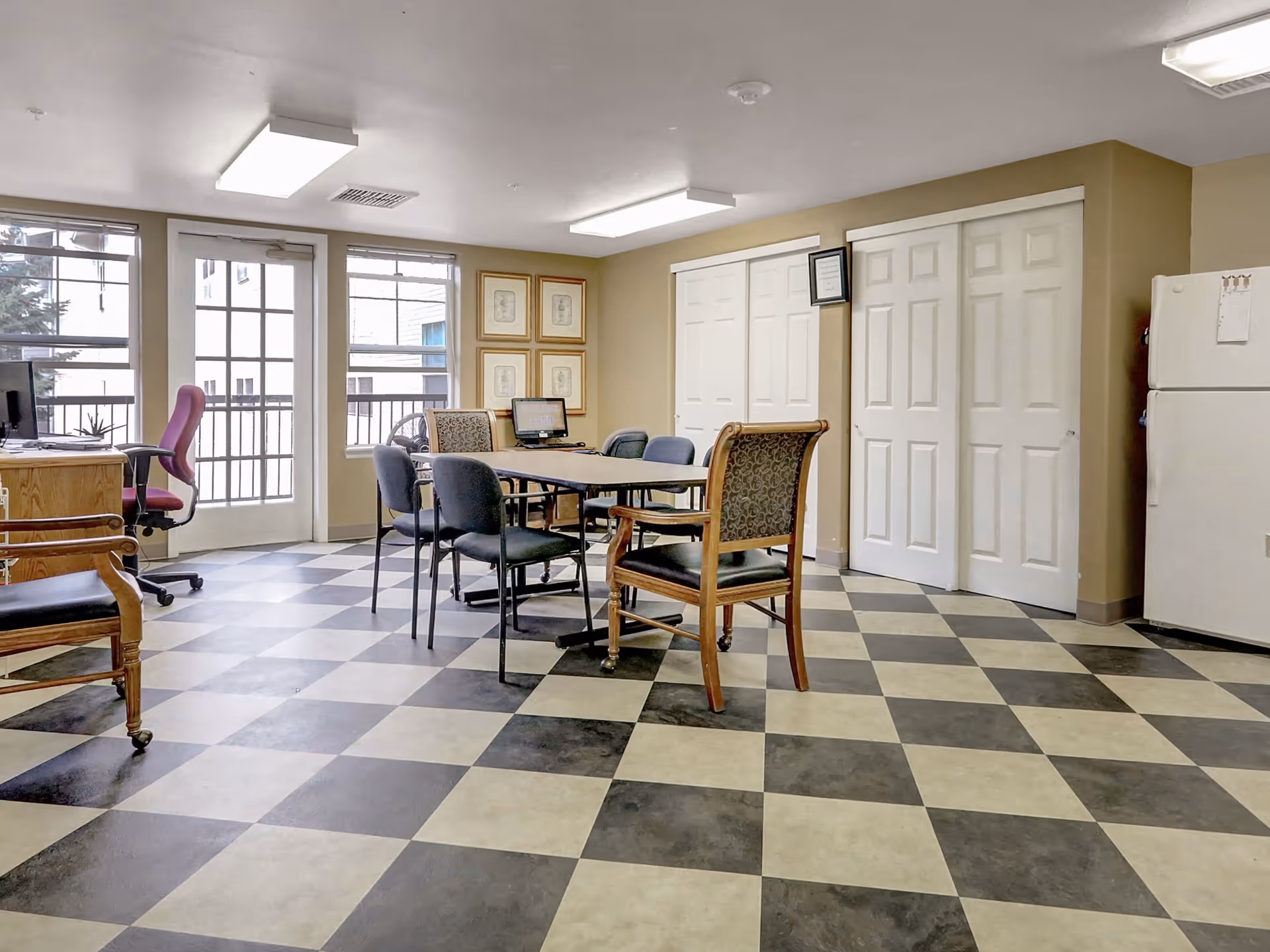 A bright room with checkered black and white flooring, featuring a rectangular table surrounded by various chairs. There is a desk with a computer and a pink office chair near large windows and a glass door leading outside. The walls are beige with framed pictures and white sliding closet doors. A white refrigerator is visible in the corner.