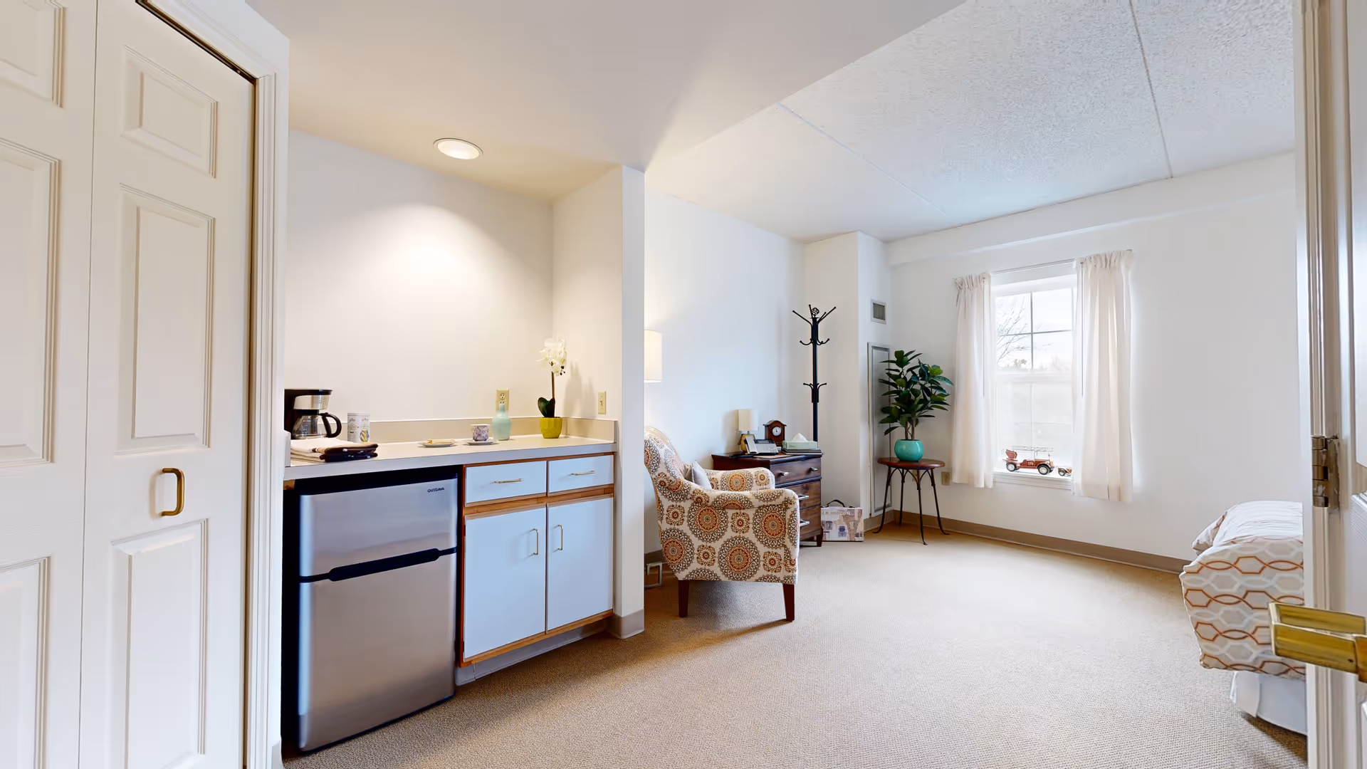 Bright studio-style living area with a kitchenette (mini fridge and cabinets), an upholstered chair, side table, window with curtains, and a partially visible bed.