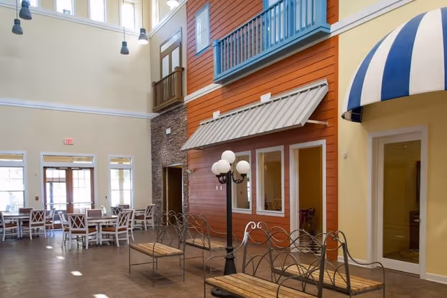 Interior view of a senior living facility common area designed to resemble a small town street with high ceilings and large windows. The space features multiple tables and chairs for dining or socializing, a lamppost with globe lights, benches with decorative metal backs, and building facades with windows, doors, awnings, and balconies painted in warm colors.