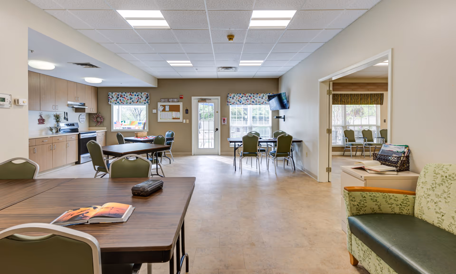A spacious, well-lit common area in a senior living facility with multiple tables and chairs arranged for group activities or dining. The room features a kitchenette with wooden cabinets, a stove, and a sink on the left side. There are windows with colorful valances, a wall-mounted TV, and a bulletin board near a door leading outside. An adjacent room with more chairs is visible through an open doorway on the right. A green patterned armchair and a small table with books are in the foreground.