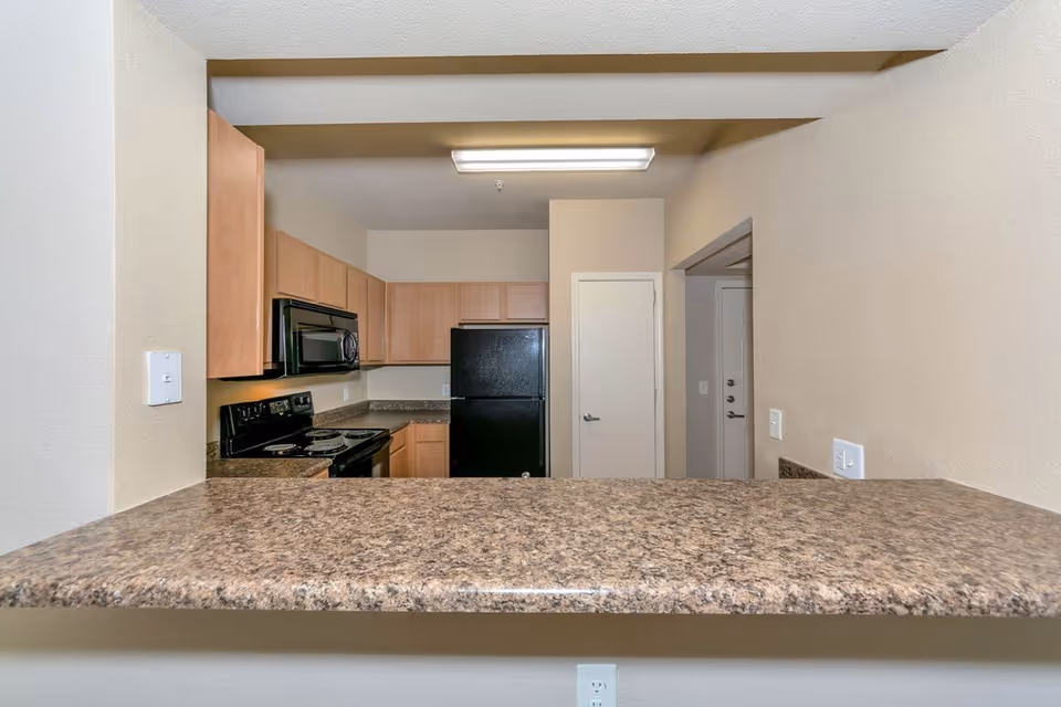 View of a kitchen area with light wood cabinets, a black refrigerator, black microwave, and black stove. The kitchen has a granite countertop bar in the foreground and beige walls.