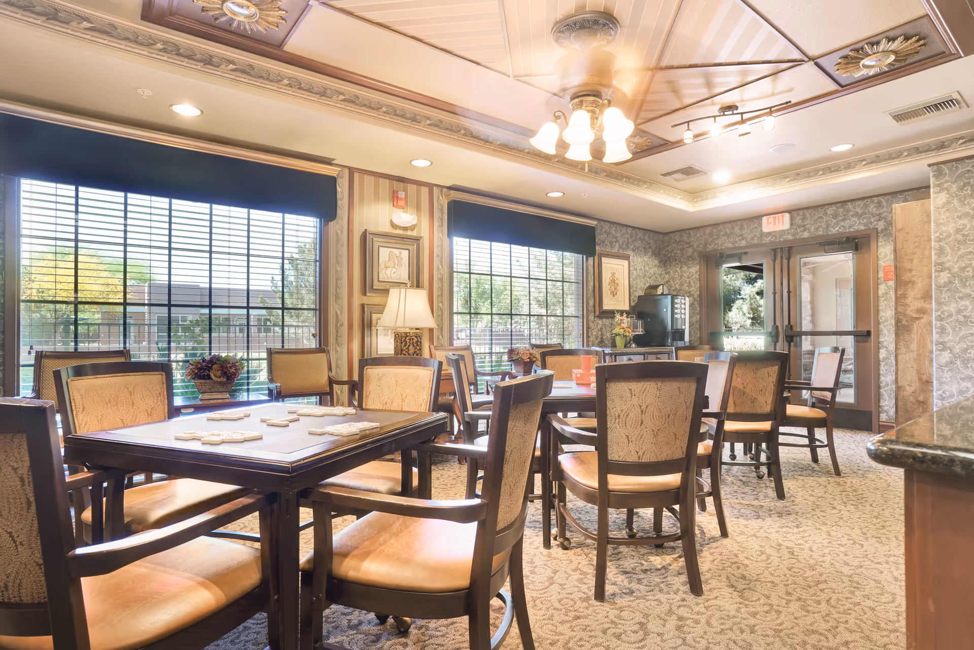 A well-lit common area with multiple tables and chairs arranged for socializing or activities. The room features large windows with blinds, decorative ceiling lights, framed artwork on the walls, and a carpeted floor. There is a coffee machine and a door leading outside in the background.