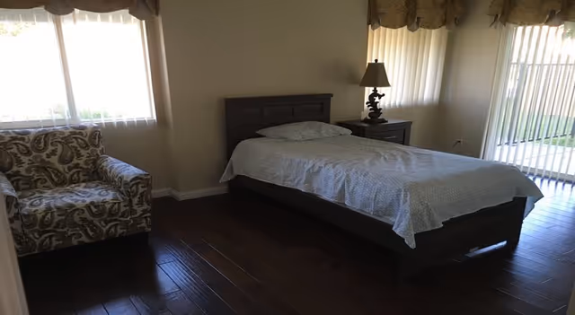 A bedroom with a single bed covered with a light-colored bedspread, a wooden nightstand with a lamp, a patterned armchair, and large windows with curtains allowing natural light to enter. The floor is dark wood, and vertical blinds cover a sliding glass door leading outside.