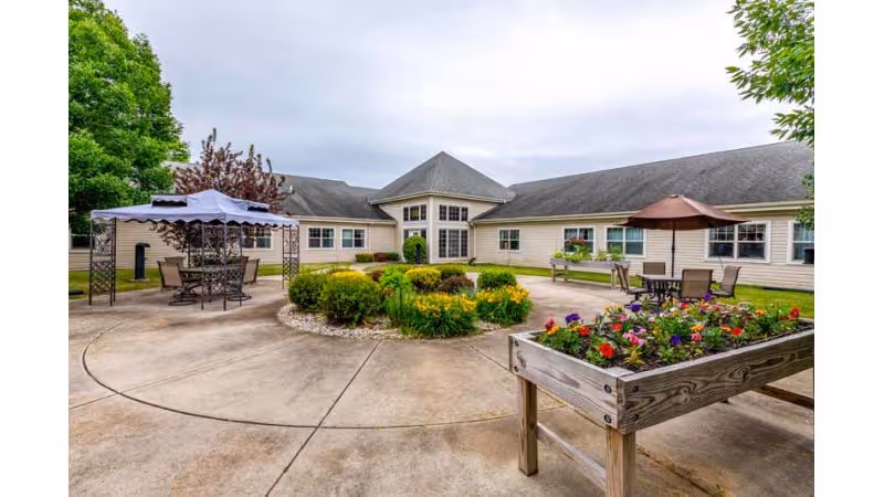 Outdoor courtyard with patio seating, umbrellas, raised flower planters and a surrounding single-story care center building.