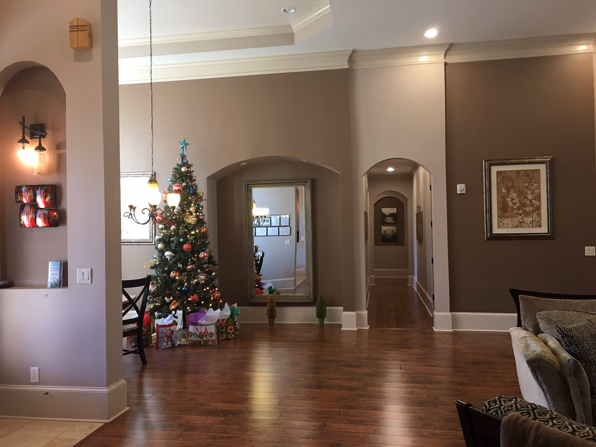 Interior view of a senior living facility decorated for Christmas with a Christmas tree adorned with ornaments and lights, surrounded by wrapped gifts. The room features wooden flooring, beige walls, a large mirror, framed artwork, and a hallway leading further into the building. There is a partial view of a dining area with chairs and a chandelier, as well as a cozy seating area with a couch and cushions.