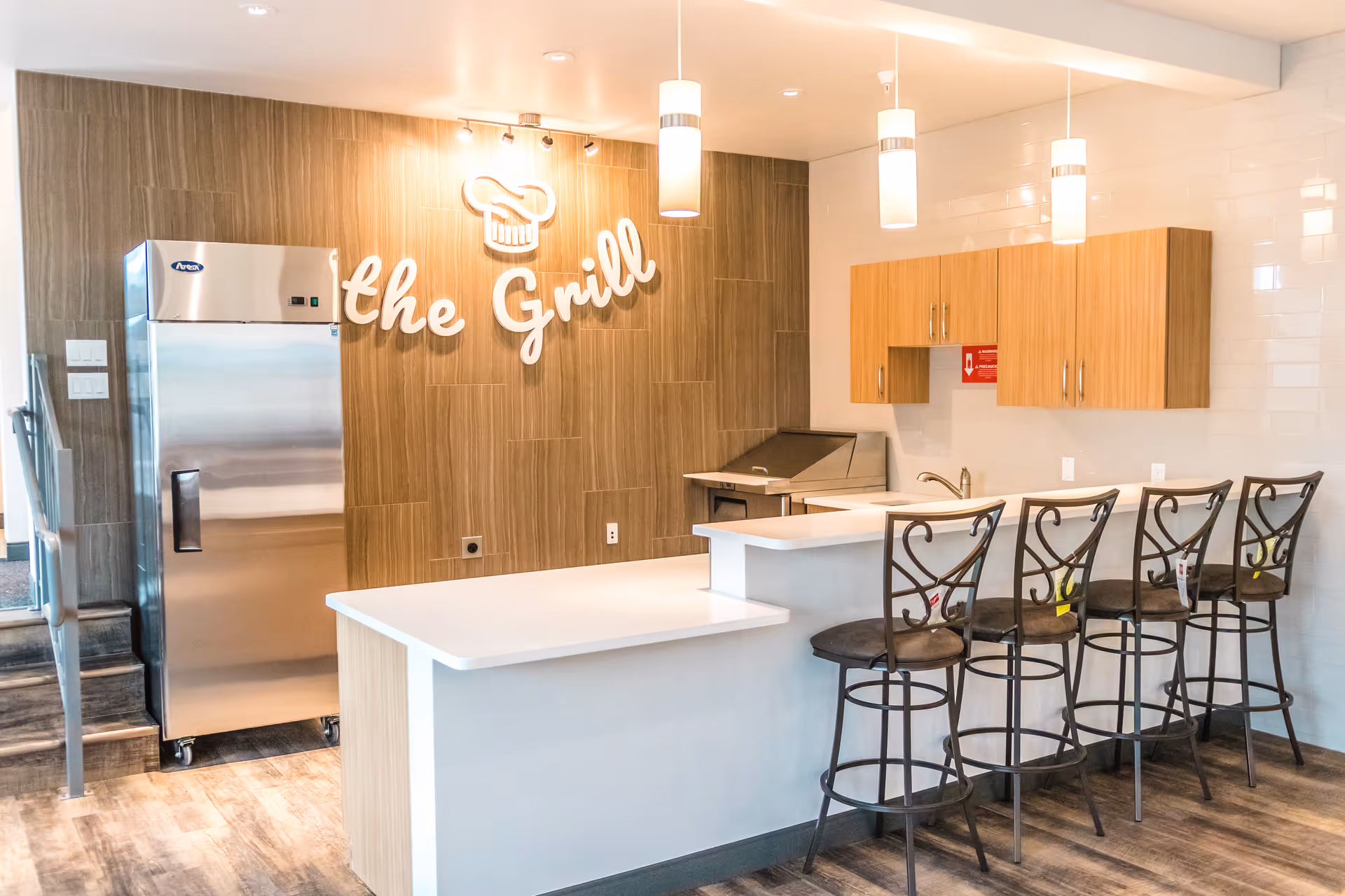Modern kitchen area with a white countertop bar and four high metal chairs with cushioned seats. Behind the bar, there are wooden cabinets mounted on a white tiled wall, a stainless steel refrigerator, and a grill station. The wall behind the grill has wood paneling with a decorative sign that reads 'the Grill' accompanied by a chef's hat icon. Three pendant lights hang from the ceiling illuminating the space.
