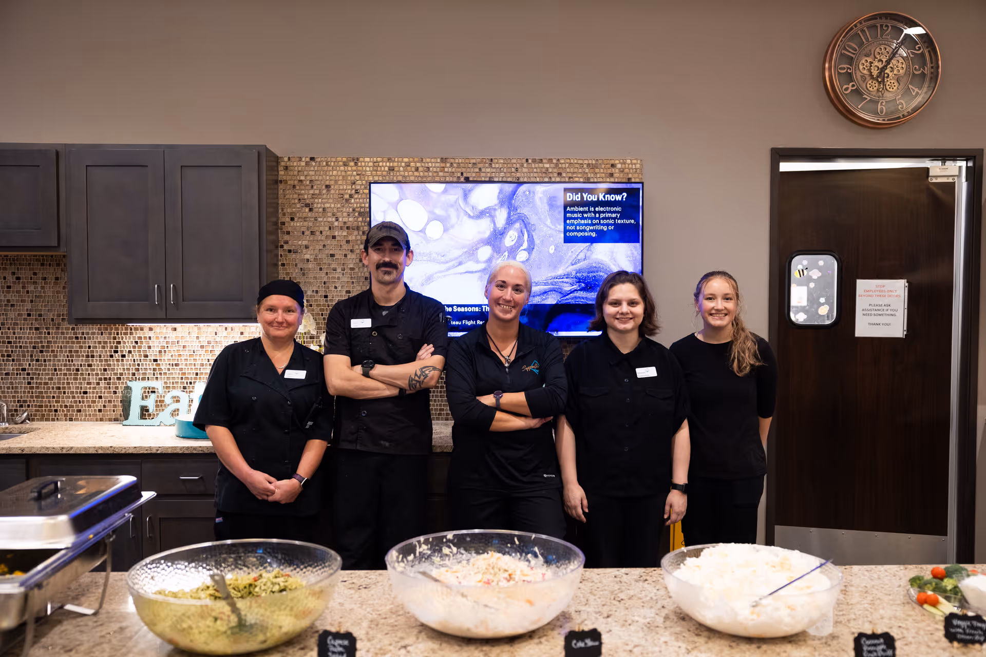 Five staff members standing behind a counter with bowls of food in front of them in a kitchen area. The staff are dressed in black uniforms and smiling. Behind them is a tiled backsplash, cabinets, a digital screen displaying information, and a door with a sign. A decorative clock is mounted on the wall above the door.