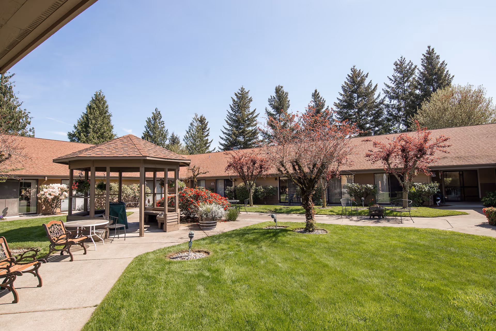 Outdoor courtyard area of a senior living facility with a gazebo, benches, tables, chairs, green grass, flowering bushes, and trees under a clear blue sky.