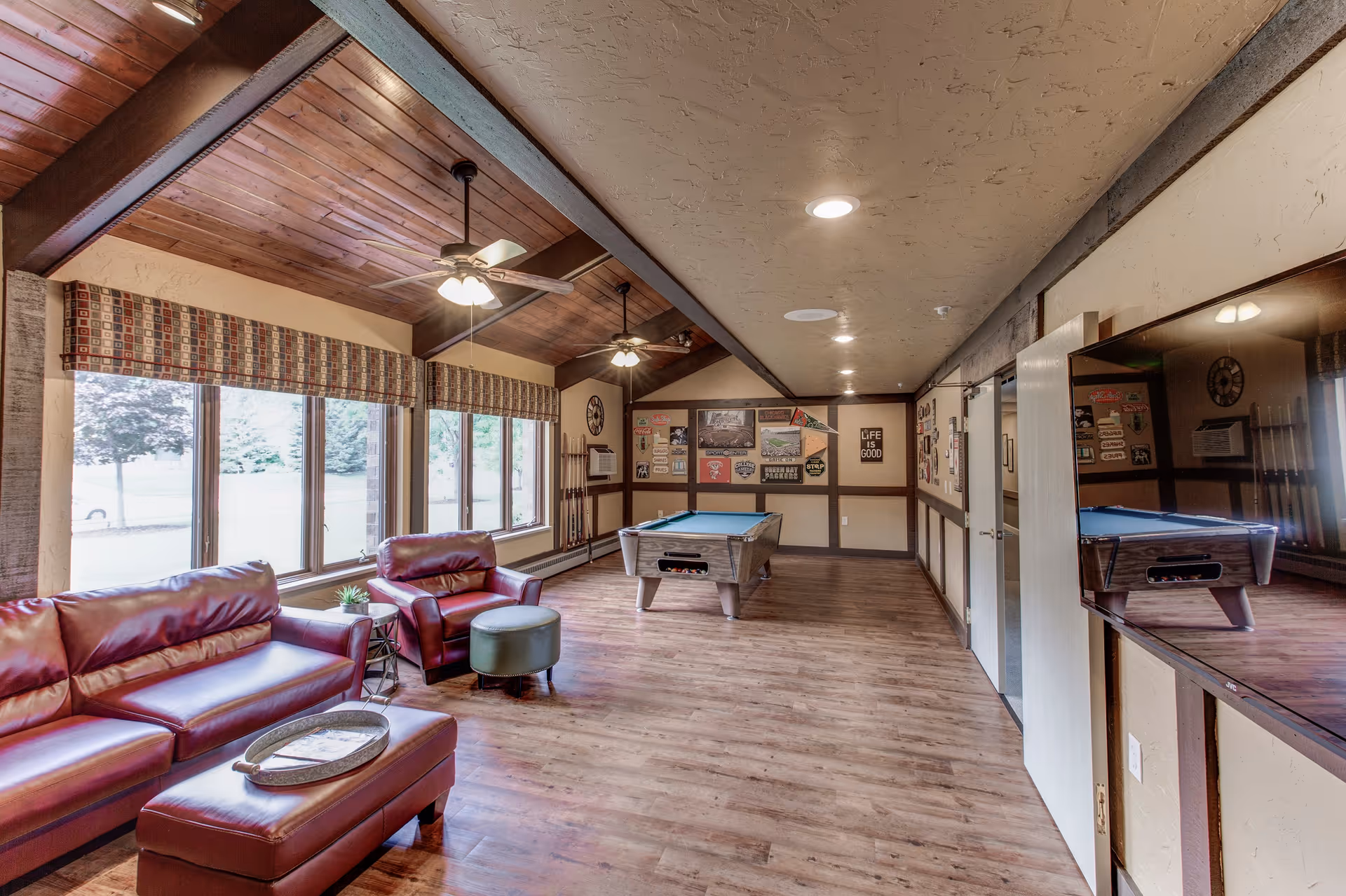 A spacious recreational room with wooden floors and a wooden ceiling with exposed beams. The room features a red leather sofa, a matching armchair with a green ottoman, and a pool table in the center. Large windows with patterned valances allow natural light to enter. The walls are decorated with various signs and posters, and a large flat-screen TV is mounted on the right wall.