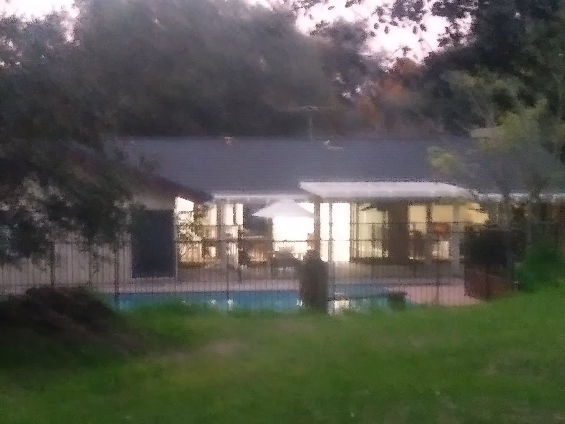 View of a single-story building with large windows illuminated from inside, surrounded by trees and greenery, with a fenced swimming pool in the foreground during dusk.