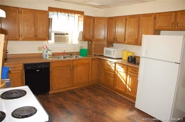 A kitchen with wooden cabinets, a white refrigerator, a white microwave, a black dishwasher, a white electric stove with coil burners, a double sink under a window with an air conditioning unit, and cleaning supplies on the counter.
