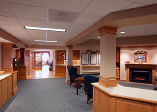 Interior view of a senior living facility common area with carpeted floors, wooden pillars, a fireplace with a framed picture above it, comfortable chairs, and a hallway leading to a bright room with large windows.