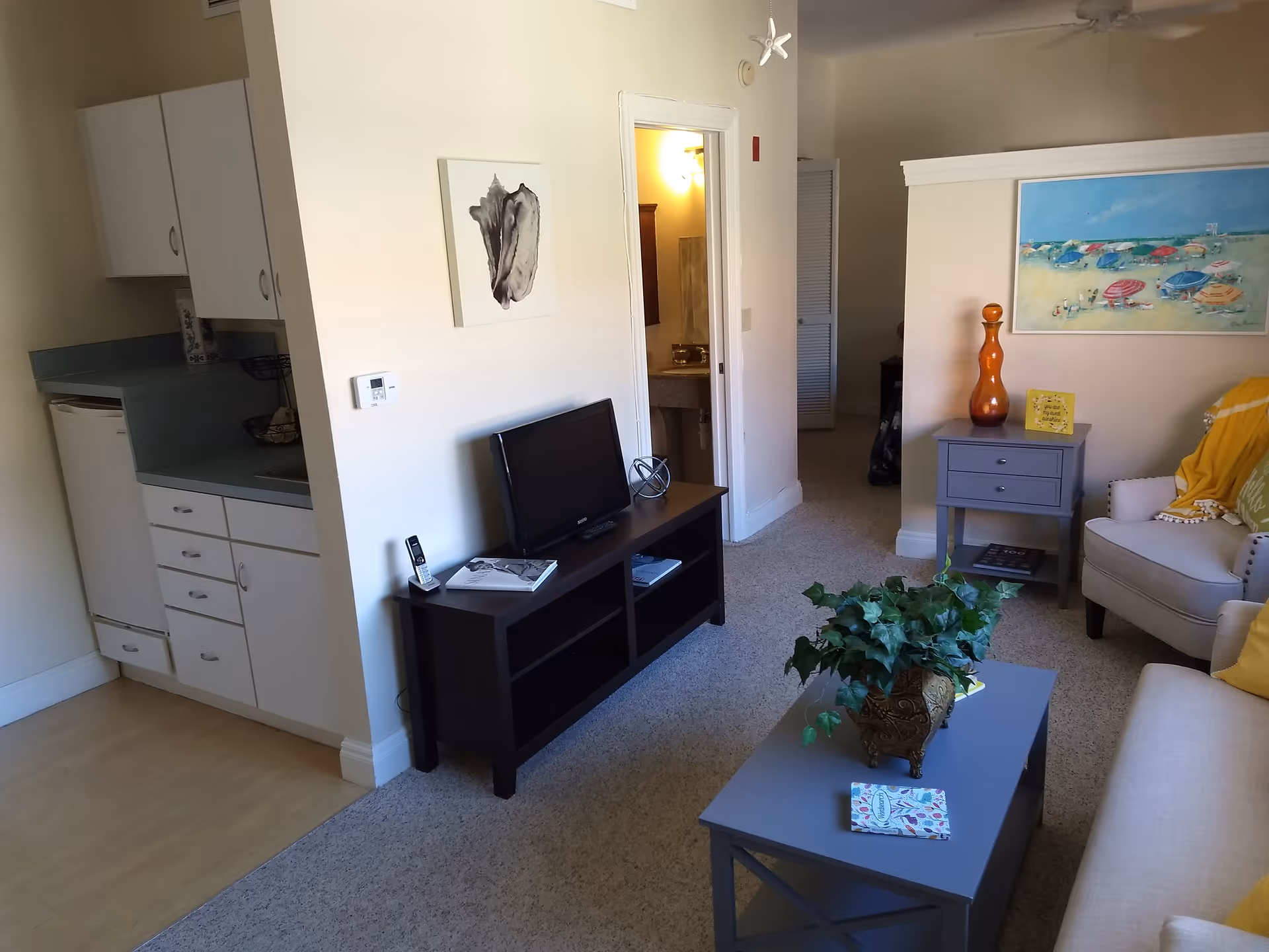 A cozy living room area with a beige sofa adorned with yellow and green pillows and a yellow throw blanket. A gray coffee table with a decorative plant and a book sits in front of the sofa. To the right, there is a gray side table with an orange vase and a small framed sign. A colorful beach-themed painting hangs on the wall above the side table. On the left side of the image, a dark wooden TV stand holds a flat-screen TV, a cordless phone, and some books. Behind the TV stand is a small kitchenette with white cabinets and a mini fridge. A doorway leads to a bathroom with a sink and mirror visible. The room has a light carpet and light-colored walls.