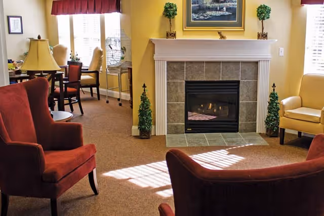 A cozy living room area with a lit fireplace surrounded by beige tiles and a white mantel. Two small decorative topiary plants flank the fireplace. There are two red upholstered armchairs and a beige armchair arranged around the fireplace. A framed painting hangs above the mantel. In the background, there is a window with red valances, a small table with chairs, and a lamp on a side table.