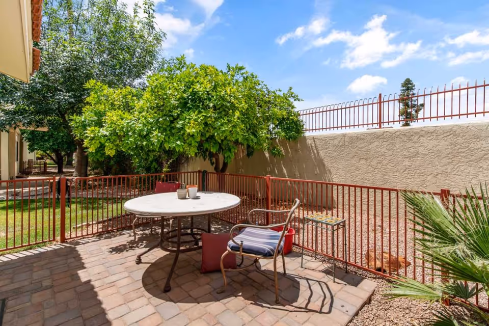 Outdoor patio area with a round table and two chairs, one with a cushion and a pillow on the ground. The patio is enclosed by a red metal fence, with trees and a grassy area visible beyond the fence. The sky is partly cloudy and blue.