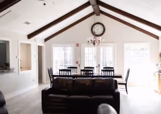 Bright dining area with a wooden table and six black chairs under a vaulted ceiling with exposed wooden beams. A large clock and chandelier hang on the wall above the table. A dark brown leather sofa is positioned in the foreground, partially blocking the view of the dining table. Large windows and glass doors allow natural light to fill the room.