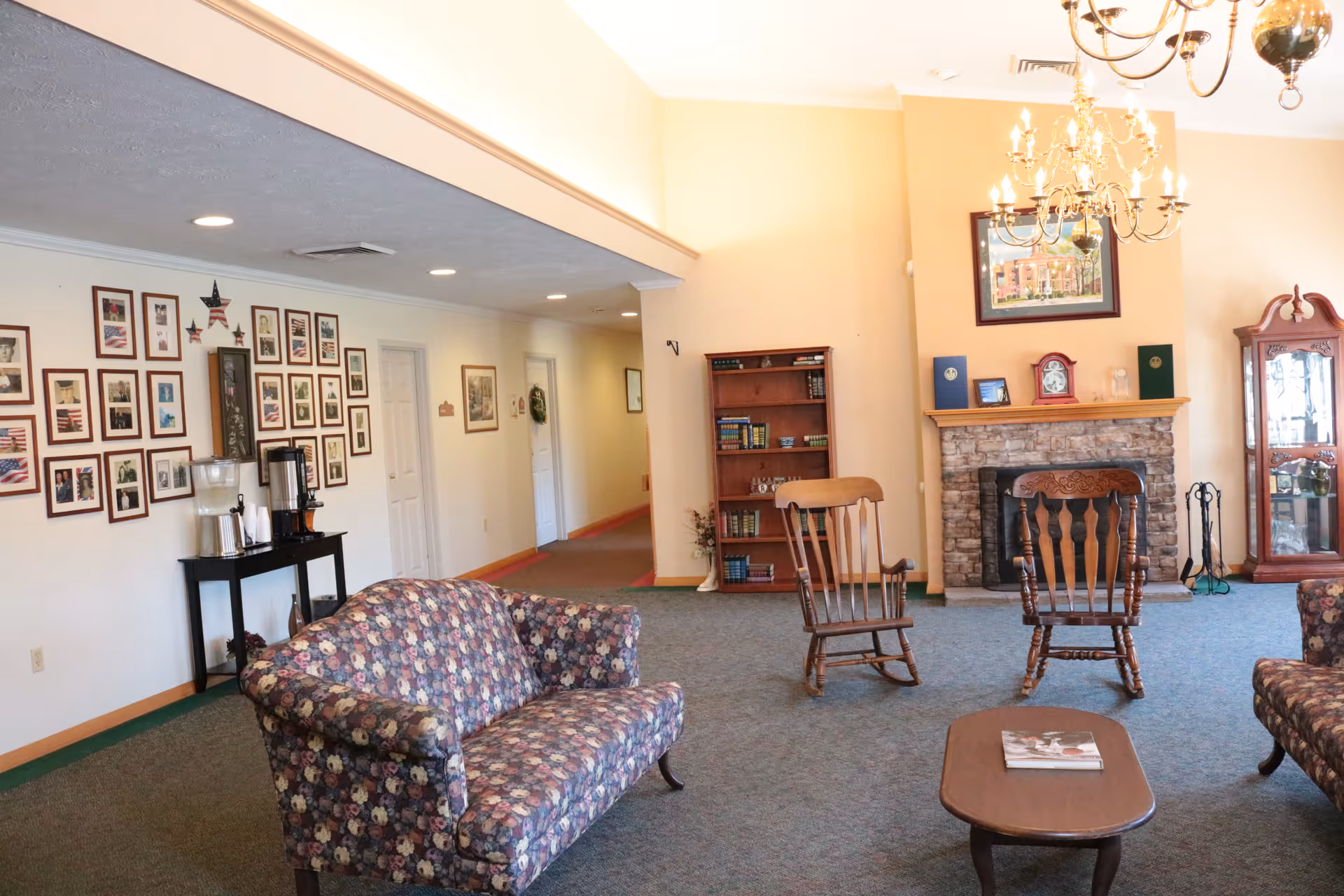 A cozy living room area in a senior living facility with floral patterned sofas, two wooden rocking chairs, a wooden coffee table with a magazine, a stone fireplace with a clock and framed picture above it, a wooden bookshelf with books, and a glass display cabinet. The walls are decorated with numerous framed photos and patriotic-themed artwork. The room is well-lit with ceiling lights and chandeliers.