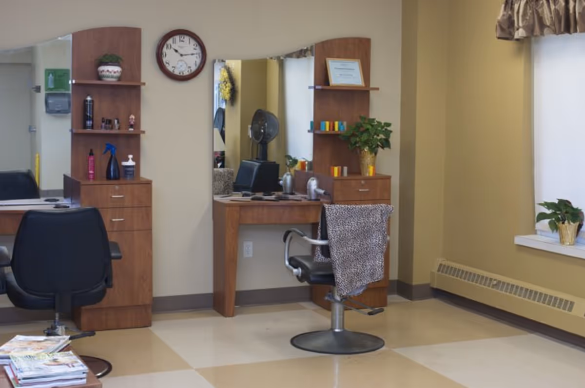 Interior view of a hair salon area in an assisted living facility with two styling stations, each with a mirror, wooden shelves, and black salon chairs. One chair has a leopard print cloth draped over it. There are various hair care products on the shelves and a clock on the wall. A window with a curtain and potted plants is visible on the right side.
