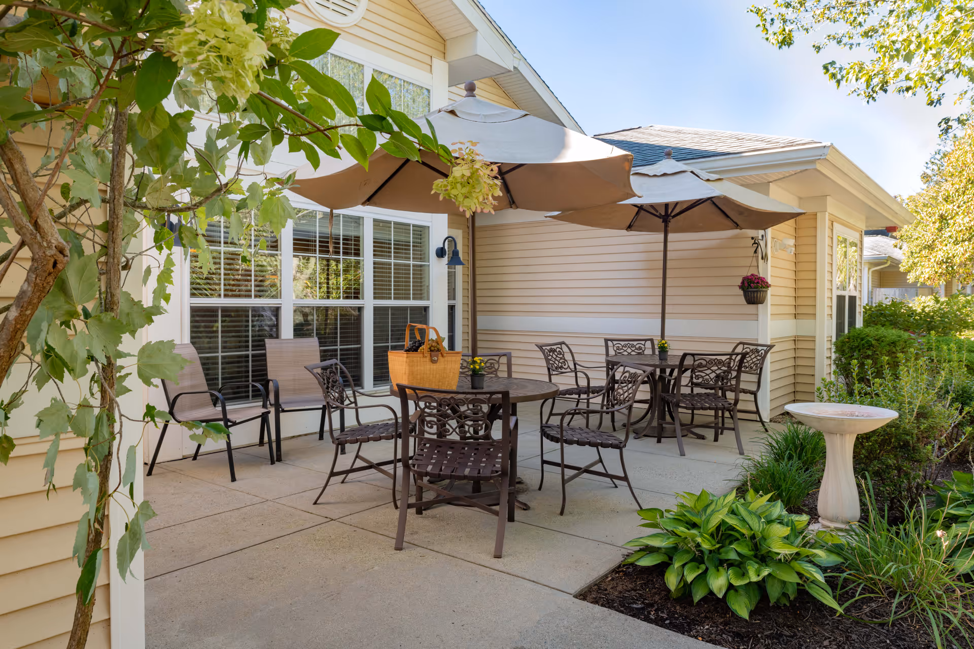 Outdoor patio with metal tables, chairs and umbrellas beside a beige siding building and surrounding greenery.