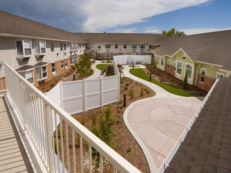 View of an outdoor courtyard area at Mountain Vista Assisted Living & Memory Care, featuring a winding concrete pathway, landscaped garden beds with small plants and trees, surrounded by a two-story building with beige and green exterior walls and multiple windows. A white fence section divides parts of the courtyard, and a balcony railing is visible in the foreground.
