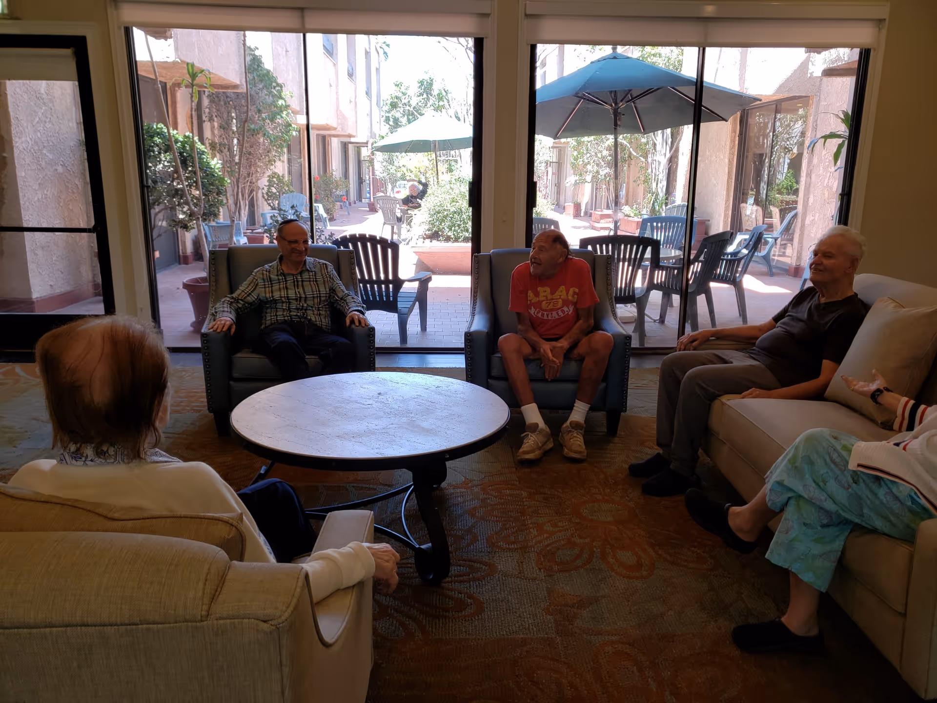 A group of elderly people sitting and chatting in a cozy indoor lounge area with large glass doors that open to an outdoor patio with tables, chairs, and umbrellas.