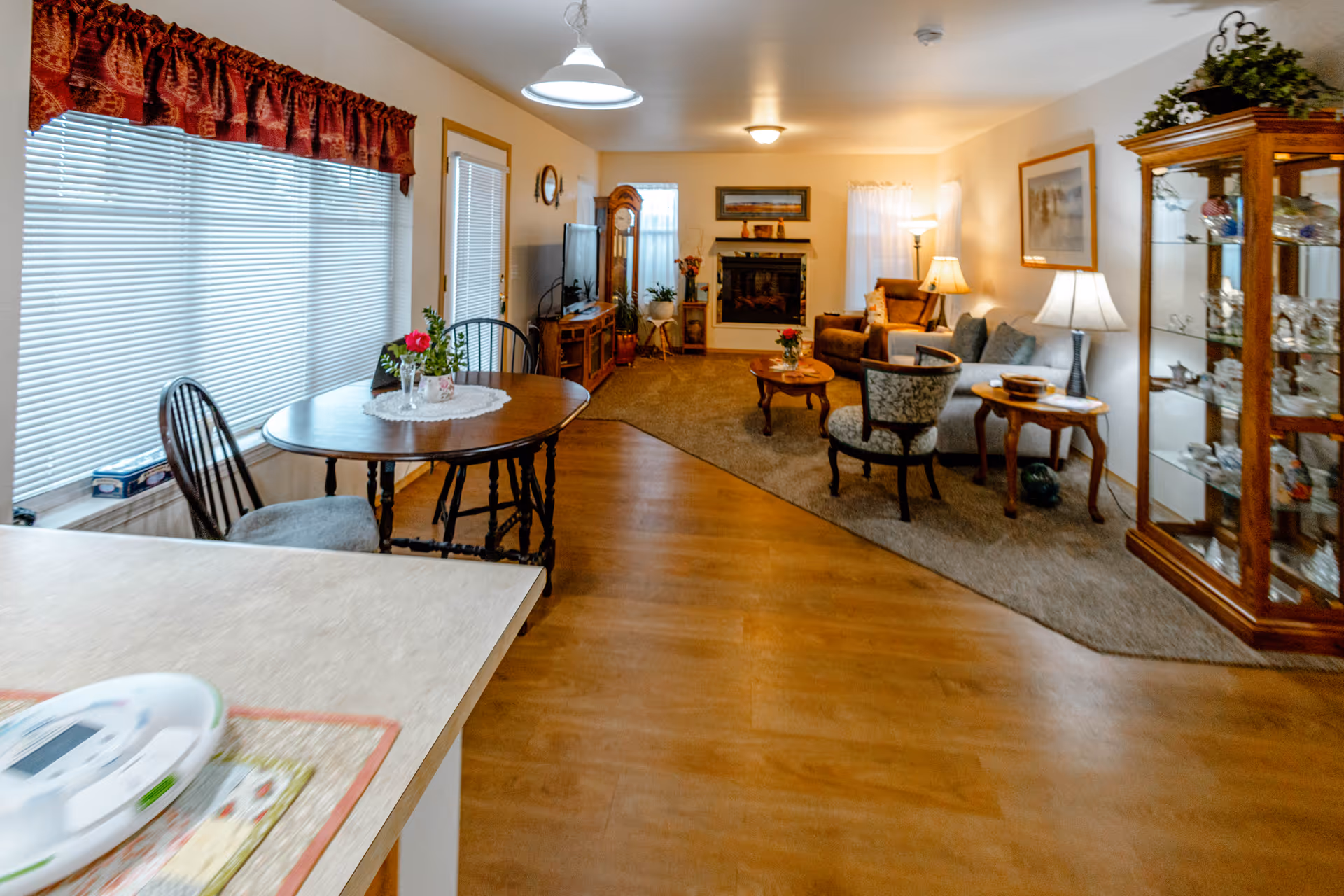 A cozy living room and dining area in a senior living facility. The room features wooden flooring, a round dining table with chairs near large windows with blinds and red valances, a carpeted seating area with armchairs, a sofa, a coffee table, side tables with lamps, a grandfather clock, a TV on a wooden stand, a fireplace, and a glass display cabinet with decorative items. The walls are decorated with framed pictures and plants add a touch of greenery.