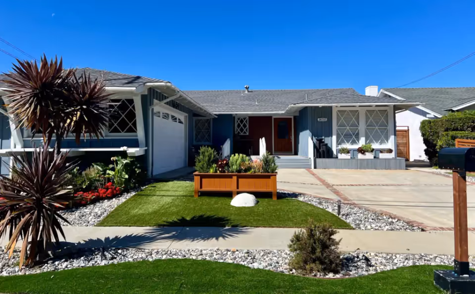 Front exterior view of a single-story house with a gray roof and blue siding. The house features a garage on the left, a front porch with steps, and large windows with white frames. The front yard has a small patch of green grass, a wooden planter box with various plants, and decorative rocks along the edges. There are also some tall spiky plants on the left side and a black mailbox on a wooden post on the right side. The sky is clear and blue.