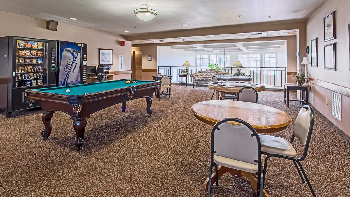 A spacious recreational room with a pool table in the center, several round tables with chairs, vending machines against the wall, and a seating area with a couch and lamps near large windows.
