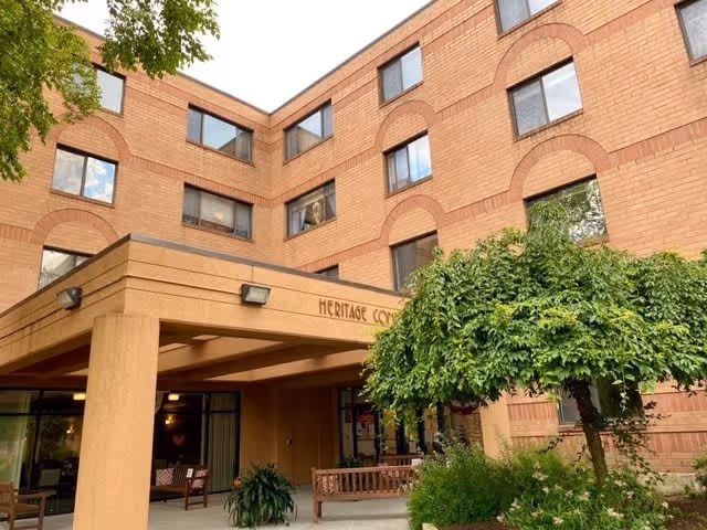 Exterior view of a multi-story brick building with large windows and a covered entrance area. There are benches and plants near the entrance, including a small tree with dense green foliage. The building has the name 'Heritage Commons' displayed above the entrance.
