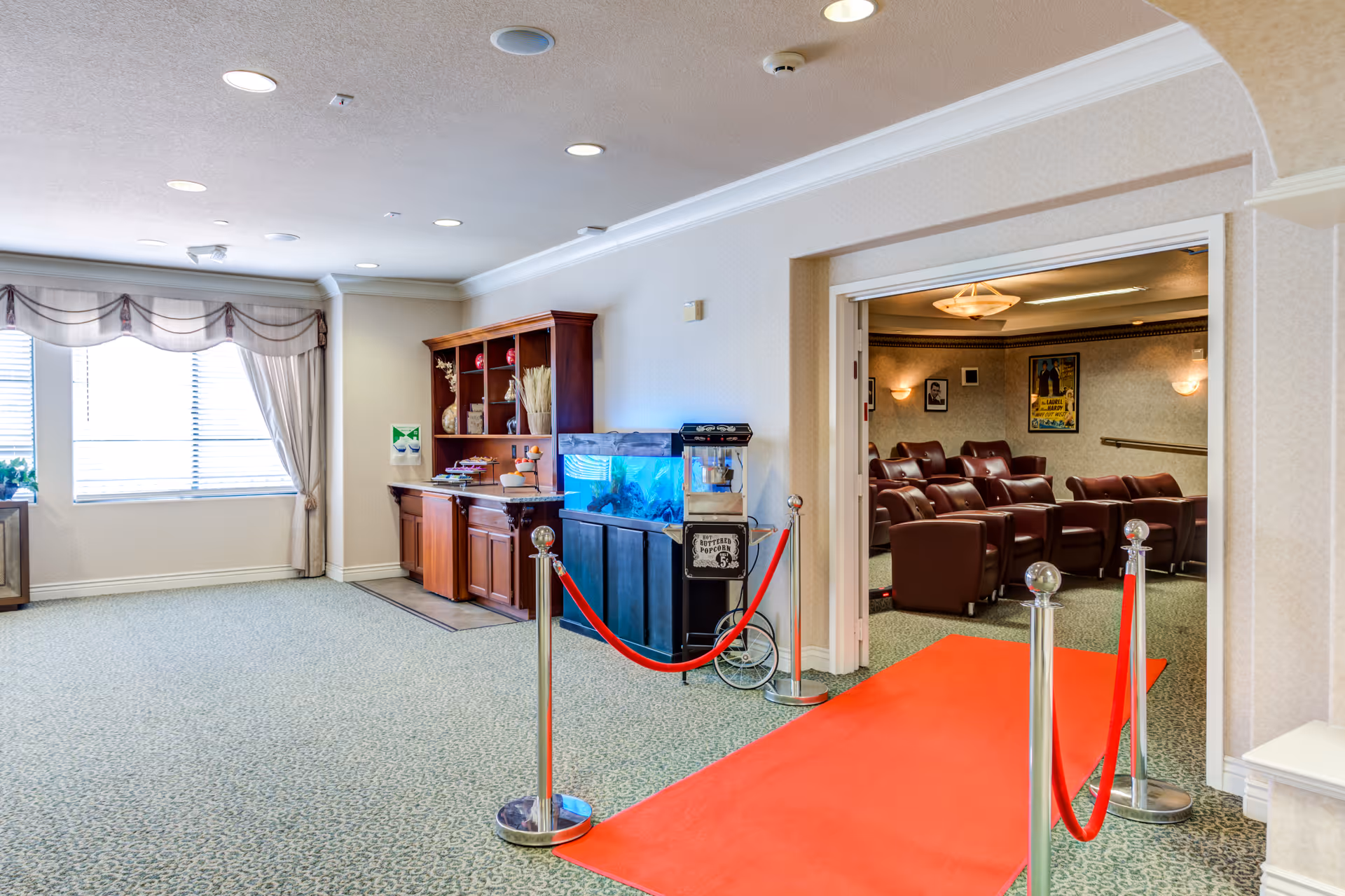 Interior view of a senior living facility showing a carpeted hallway with a red carpet leading to a small theater room with brown leather recliners. To the left, there is a wooden cabinet with shelves and an aquarium on top, along with a popcorn machine. Large windows with curtains allow natural light into the space.