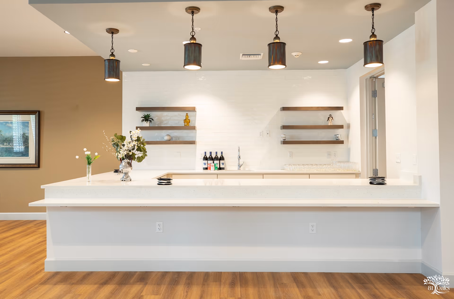 Communal kitchen/bar area with a long white counter, pendant lights, floating shelves, and decorative bottles and flowers.