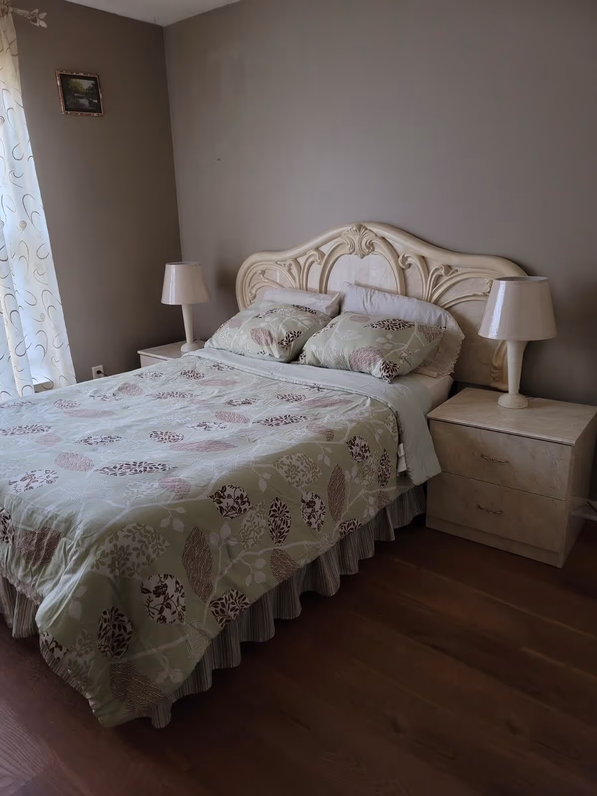 A bedroom with a double bed featuring a decorative cream-colored headboard. The bed is made with a green and beige patterned bedspread and matching pillows. There are two beige bedside tables, each with a matching lamp. A window with sheer white curtains is visible on the left side, and a small framed picture hangs on the wall above the bed.