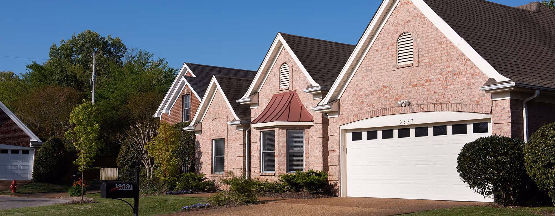 Exterior view of a brick residential building with a white garage door, multiple gabled roofs, and well-maintained landscaping including bushes and trees under a clear blue sky.