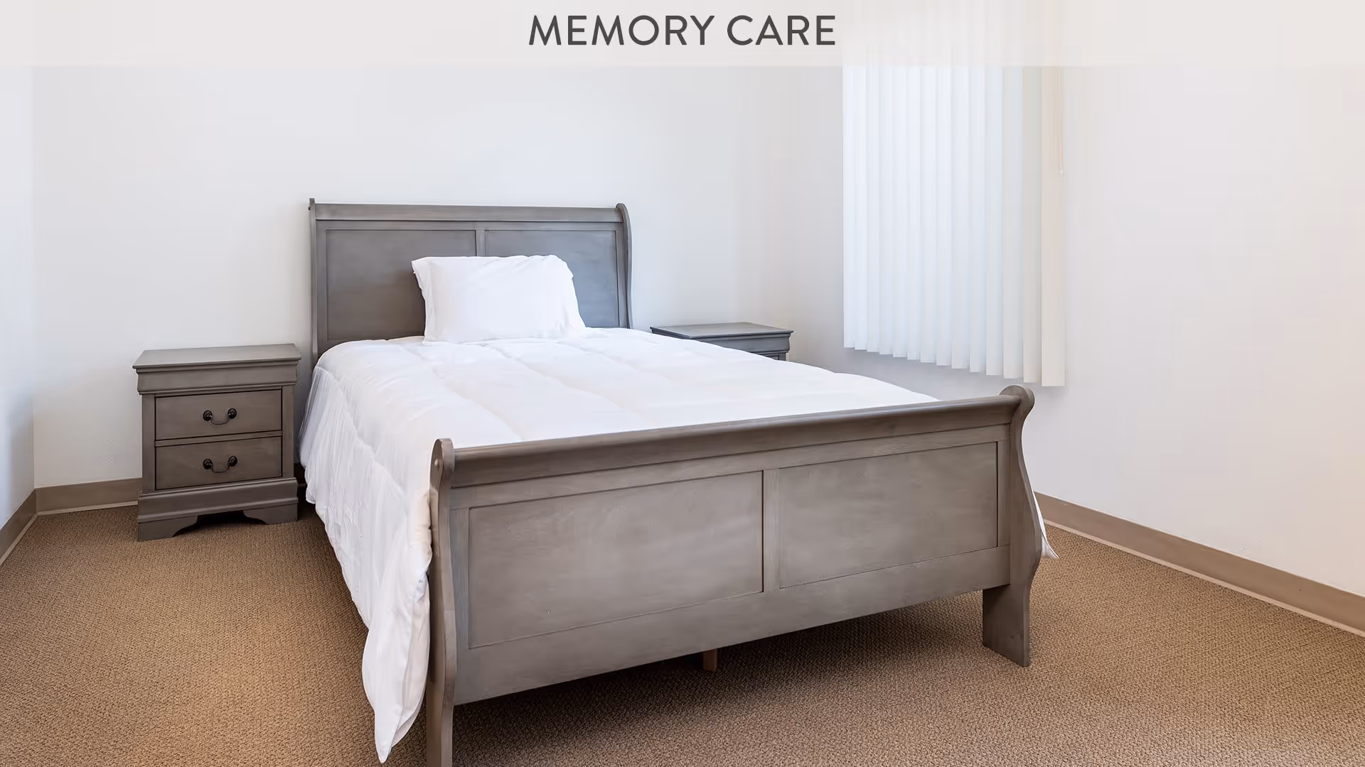 A simple bedroom with a gray wooden bed frame and white bedding, flanked by two matching gray nightstands with drawers, beige carpet flooring, white walls, and a window with vertical blinds.