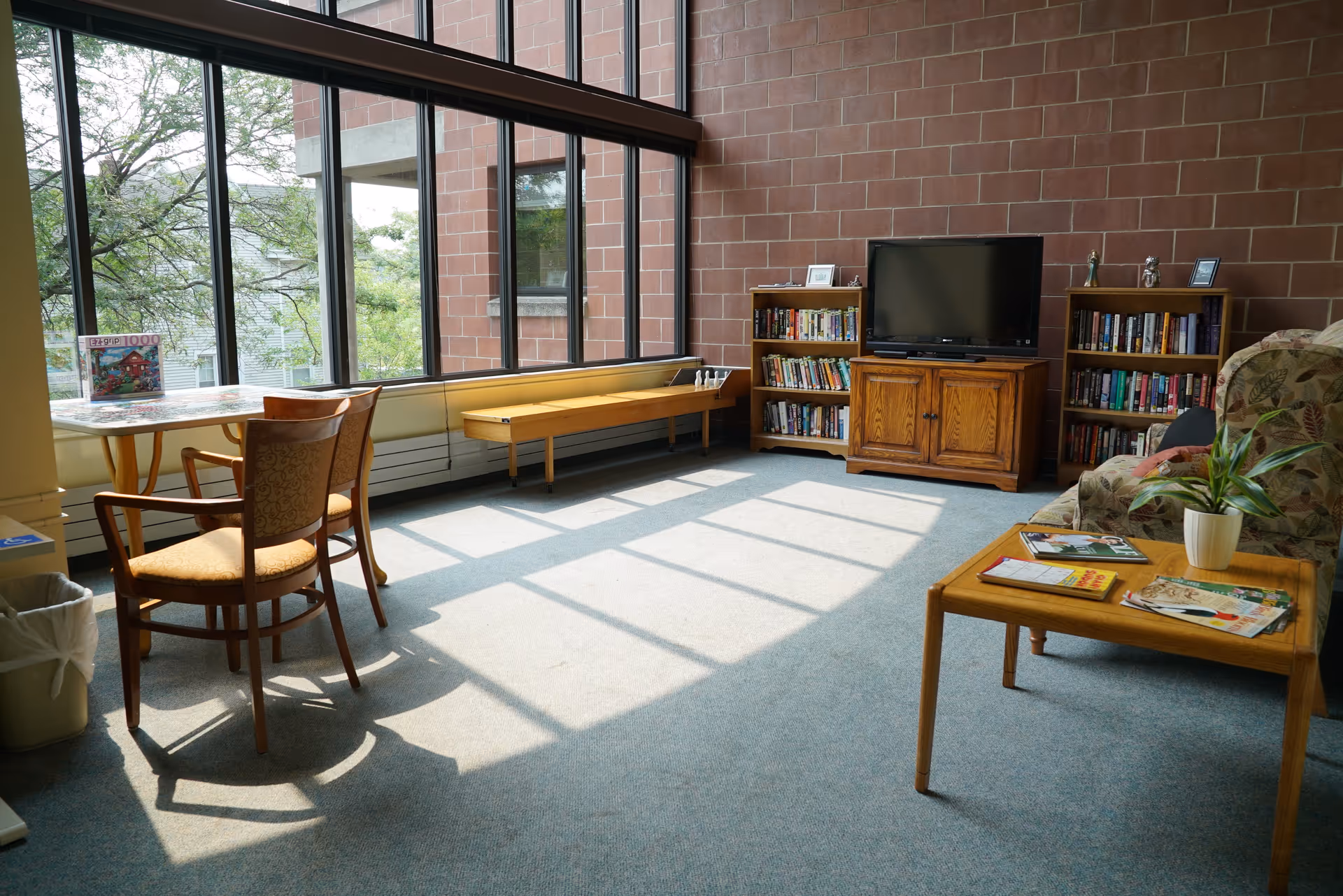 A bright common area with large windows letting in natural light, featuring a small table with two chairs, a wooden shuffleboard game, a TV on a wooden cabinet flanked by two bookshelves filled with books, and a floral-patterned armchair next to a wooden coffee table with magazines and a potted plant.