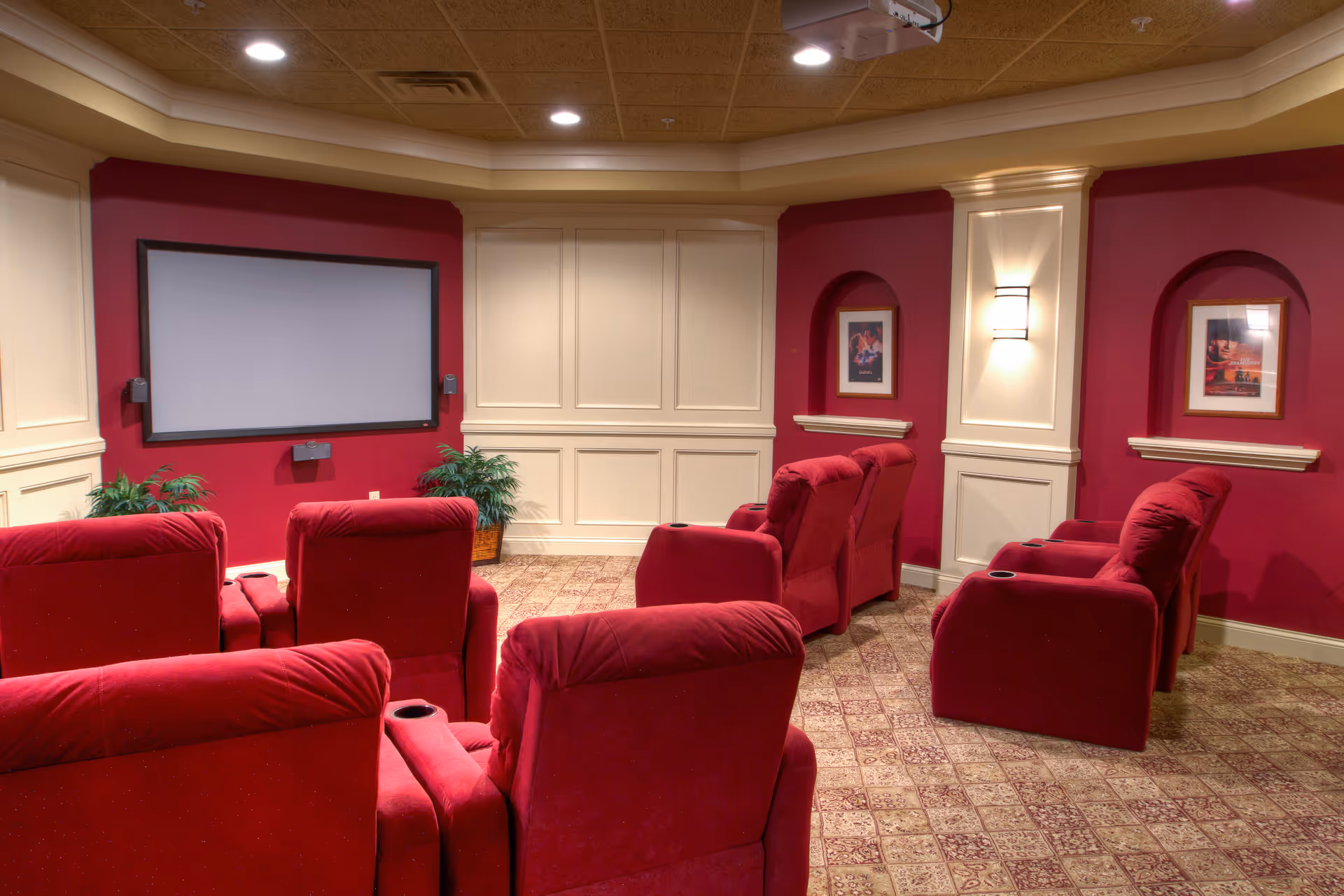 A cozy senior living facility theater room with red upholstered reclining chairs arranged in rows facing a large blank projection screen. The walls are painted red with cream paneling and decorative niches holding framed pictures. The carpet has a patterned design, and there are ceiling lights and a projector mounted above.