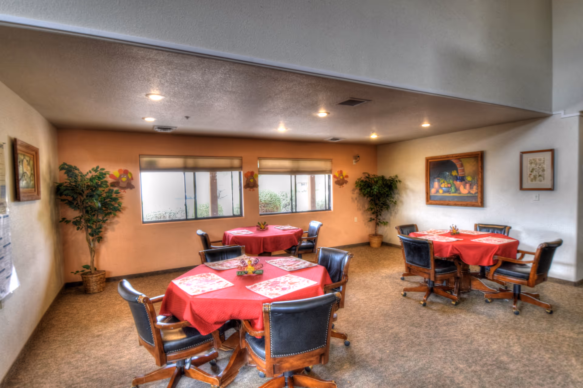 Communal dining room with round tables draped in red tablecloths, rolling wooden chairs, potted plants, wall art, and windows letting in natural light.
