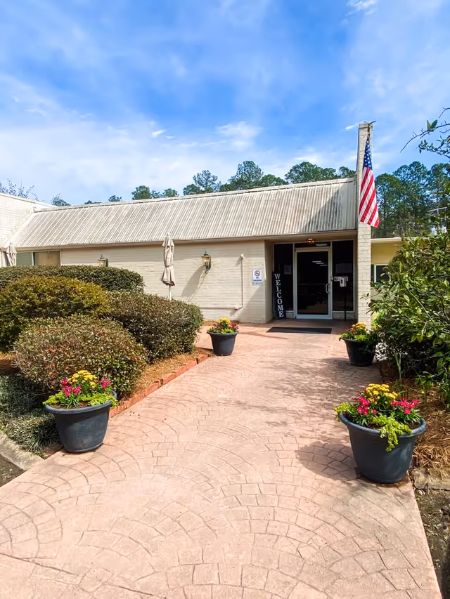 Entrance walkway to a single-story building with a beige exterior and a slanted roof. The walkway is lined with four large flower pots containing colorful flowers and greenery. There are trimmed bushes on the left side and an American flag mounted on a pillar near the entrance. A vertical 'WELCOME' sign is visible next to the glass double doors under a clear blue sky.