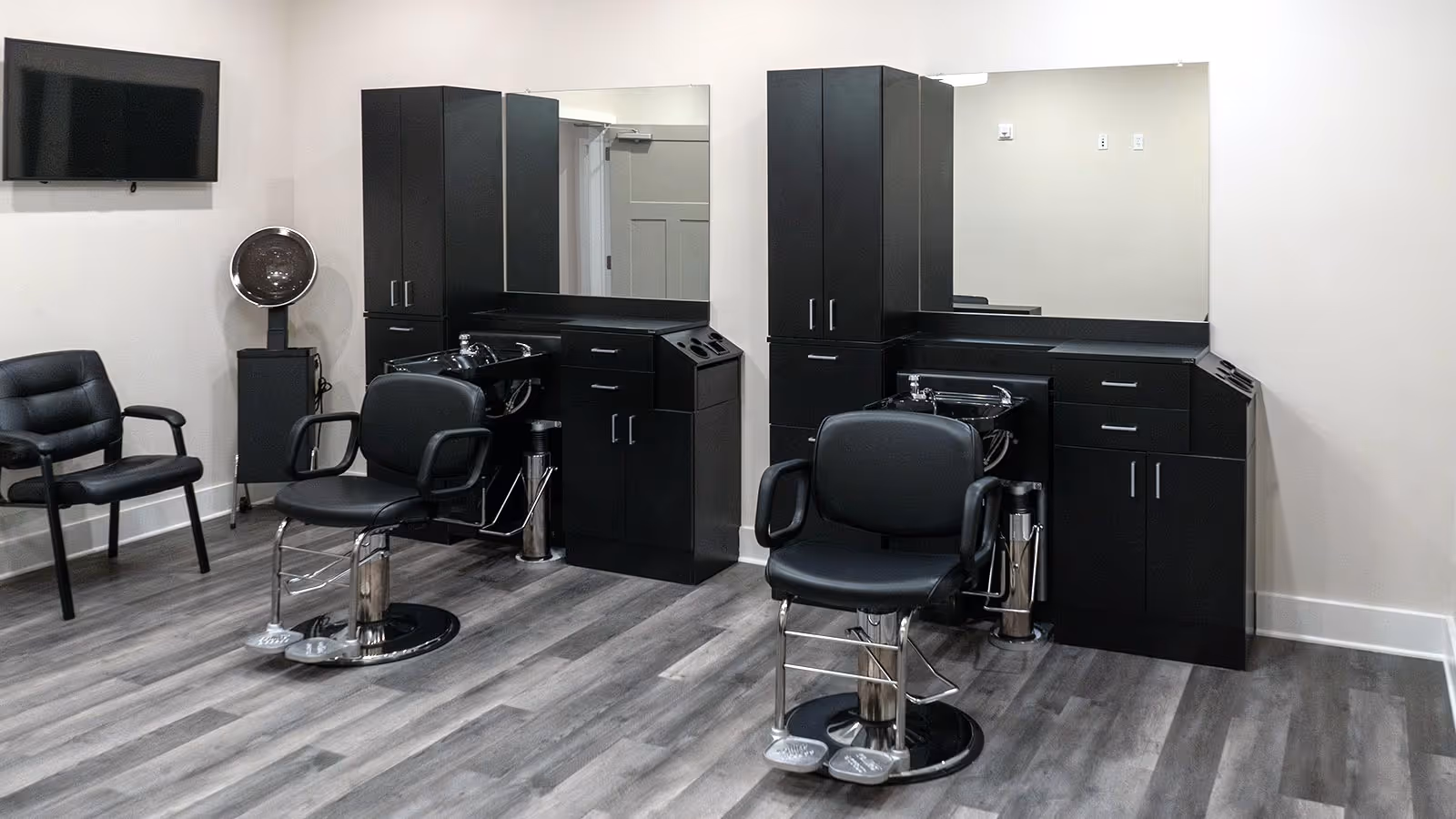 Salon-style grooming area with two styling chairs, shampoo bowls, large mirrors, black cabinets and a hooded hair dryer.