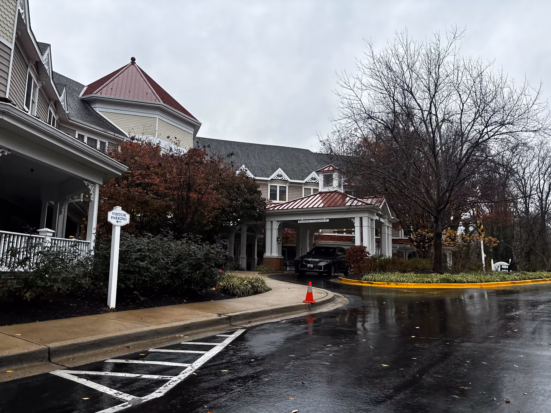 Exterior view of Kensington Park Senior Living facility on a cloudy day, showing the entrance with a covered drop-off area, a black vehicle parked under the canopy, a visitor parking sign, leafless trees, and wet pavement.