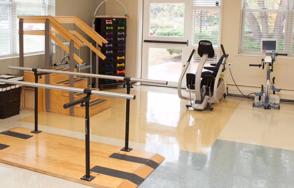 A rehabilitation or physical therapy room with parallel bars for walking exercises, a set of stairs with handrails, a rack of colorful dumbbells, and two exercise machines near windows with blinds.